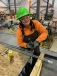 woman wearing safety glasses and a hard hat smiles while holding a drill at a work station